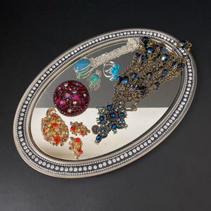 Puede incluir: Una bandeja ovalada de plata con una superficie de espejo y un borde de cuentas. La bandeja está llena de varias piezas de joyería, incluyendo un gran broche rojo, un broche azul y dorado, un colgante turquesa, un anillo de plata y un largo collar de cuentas.