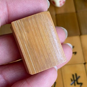 May include: A close-up of a light brown rectangular Mahjong tile. The tile is made of wood and has a textured surface with visible grain. The tile is held in a person's hand. Other Mahjong tiles and a die are visible in the background.