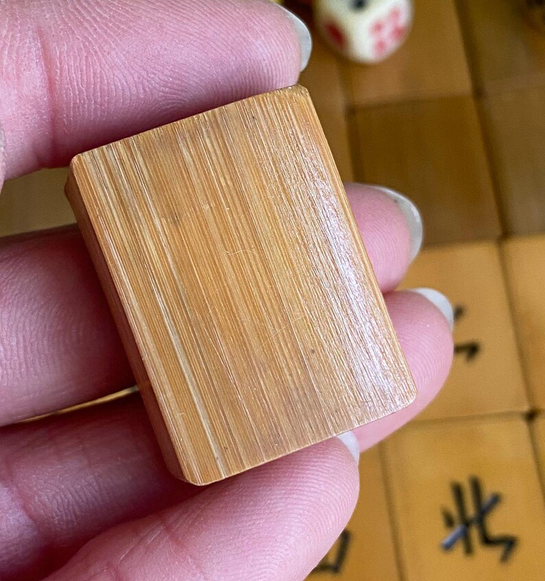 May include: A close-up of a light brown rectangular Mahjong tile. The tile is made of wood and has a textured surface with visible grain. The tile is held in a person's hand. Other Mahjong tiles and a die are visible in the background.