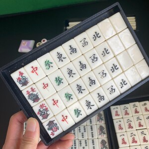 May include: A set of ivory-colored Mahjong tiles with black, red, and green symbols and characters. The tiles are arranged in a black tray, with additional tiles visible in the background. The game pieces are for the traditional Chinese tile-based game.