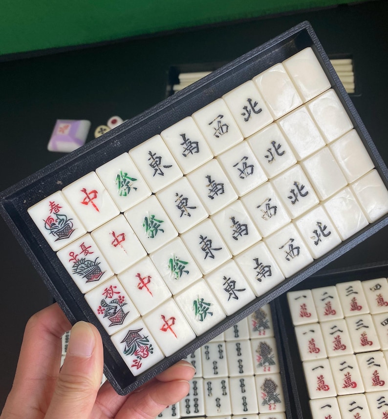 May include: A set of ivory-colored Mahjong tiles with black, red, and green symbols and characters. The tiles are arranged in a black tray, with additional tiles visible in the background. The game pieces are for the traditional Chinese tile-based game.