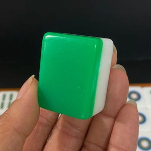 May include: A close-up of a green and white Mahjong tile. The tile is rectangular with a glossy green surface on one side and a white surface on the other. The tile is held in a person's hand.