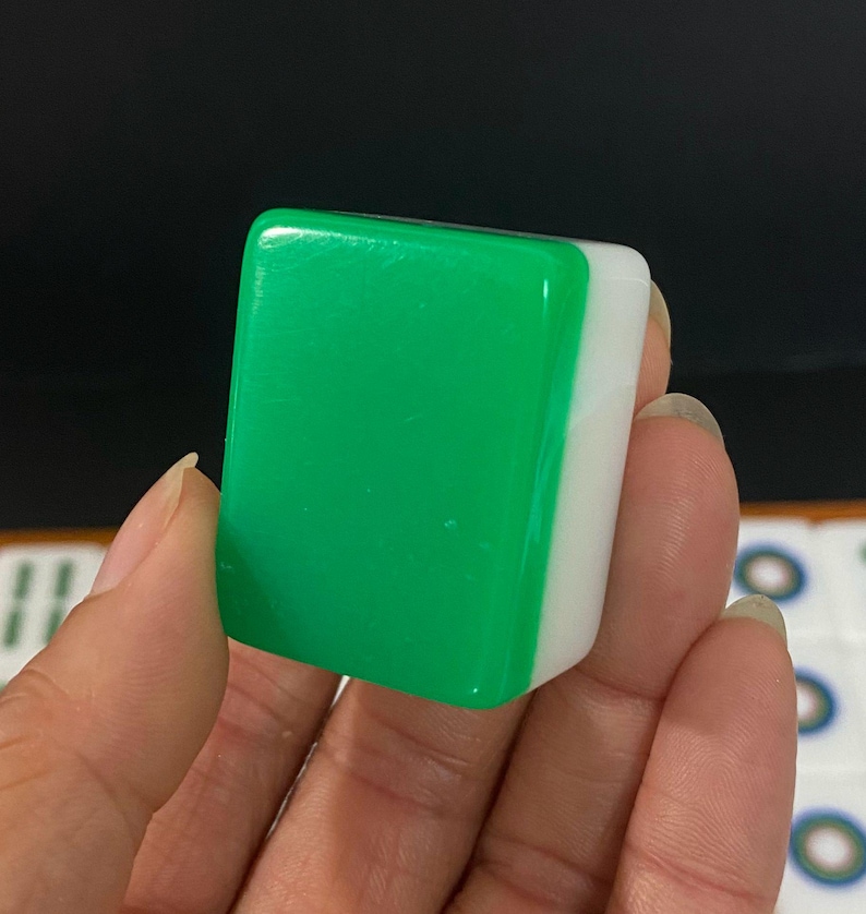 May include: A close-up of a green and white Mahjong tile. The tile is rectangular with a glossy green surface on one side and a white surface on the other. The tile is held in a person's hand.