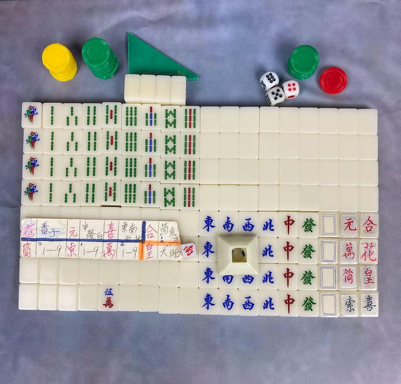 May include: A complete Mahjong set with ivory-colored tiles, dice, and chips. The tiles feature Chinese characters and symbols in red, green, and blue. Includes a green triangular piece and various colored chips.
