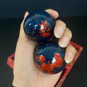Puede incluir: Dos bolas decorativas azules con detalles rojos y dorados. Las bolas tienen un aspecto veteado y presentan un diseño floral. Se sostienen en una mano y descansan en una caja roja.