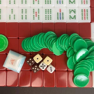 May include: A complete Mahjong set with white tiles featuring green, red, and blue markings. The set includes green chips, dice, and a small, square, blue and white container. The tiles are arranged on a red surface.