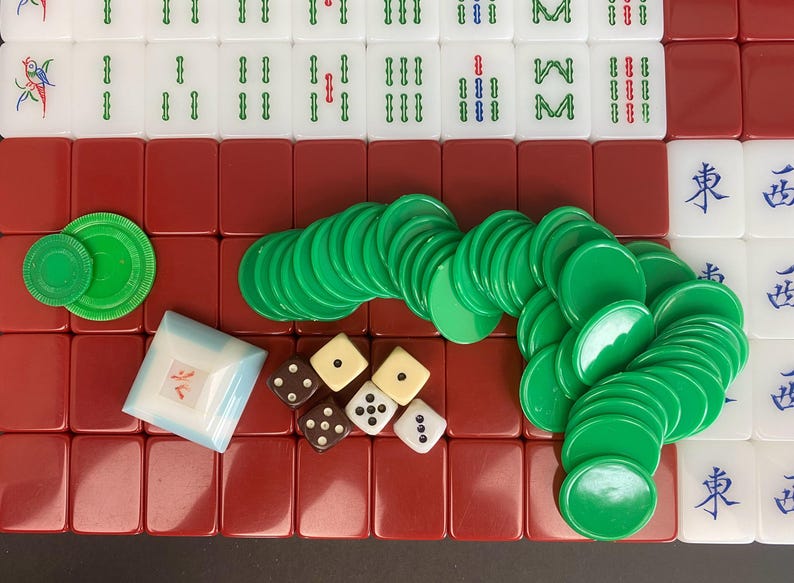 May include: A complete Mahjong set with white tiles featuring green, red, and blue markings. The set includes green chips, dice, and a small, square, blue and white container. The tiles are arranged on a red surface.