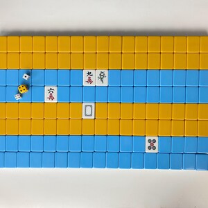 Puede incluir: Disposici&oacute;n rectangular de fichas de Mahjong amarillas y azules. Varias fichas muestran caracteres y s&iacute;mbolos chinos. Dados con caras blancas y amarillas est&aacute;n dispersos cerca de las fichas, lo que sugiere un juego en curso. Las fichas est&aacute;n dispuestas sobre una superficie blanca.