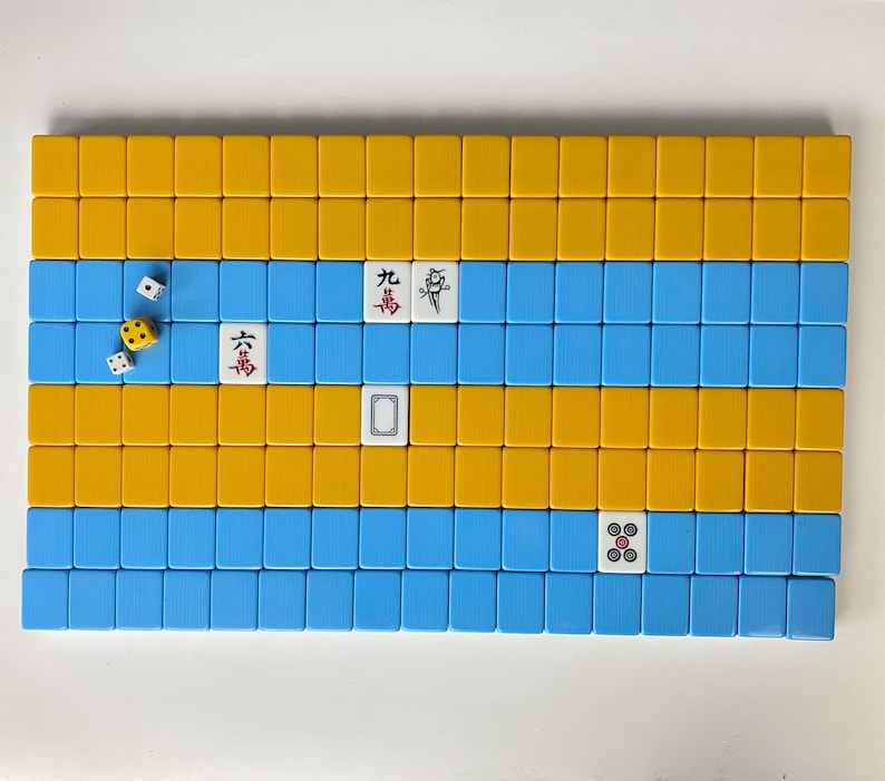 Puede incluir: Disposici&oacute;n rectangular de fichas de Mahjong amarillas y azules. Varias fichas muestran caracteres y s&iacute;mbolos chinos. Dados con caras blancas y amarillas est&aacute;n dispersos cerca de las fichas, lo que sugiere un juego en curso. Las fichas est&aacute;n dispuestas sobre una superficie blanca.