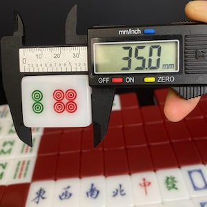May include: A white Mahjong tile with green and red circular designs is measured with a digital caliper. The caliper displays 35.0 mm. Other Mahjong tiles with Chinese characters are visible in the background.
