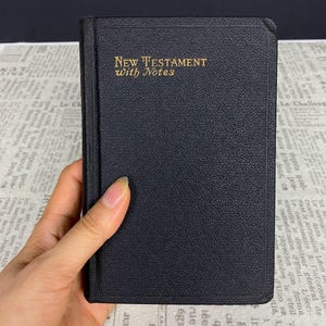 May include: A black New Testament with gold lettering reading "New Testament with Notes." The book is held in a hand. The background is printed paper.
