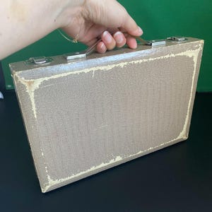 May include: A vintage, rectangular suitcase with a textured, faux-leather exterior in a muted taupe color. The suitcase has a silver metal handle and clasps, and the edges show signs of wear. The overall aesthetic suggests a classic, travel-themed item.