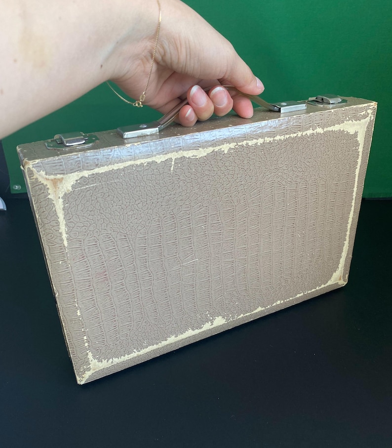 May include: A vintage, rectangular suitcase with a textured, faux-leather exterior in a muted taupe color. The suitcase has a silver metal handle and clasps, and the edges show signs of wear. The overall aesthetic suggests a classic, travel-themed item.