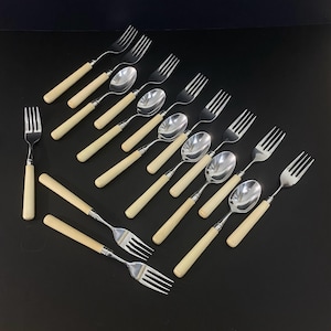 May include: A set of stainless steel cutlery comprising several forks and spoons, each featuring a smooth, cream-coloured handle. The utensils are arranged in a diagonal pattern, showcasing the metallic heads and the uniform light-coloured handles.