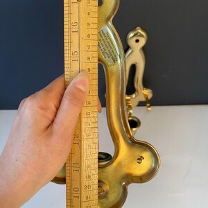 May include: A gold-colored wall sconce with a curved design, approximately 11 inches tall. The sconce has a decorative shape and a small cup for holding a candle. A wooden ruler is held next to the sconce.
