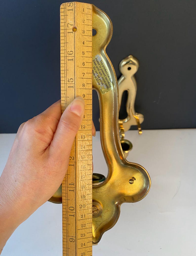 May include: A gold-colored wall sconce with a curved design, approximately 11 inches tall. The sconce has a decorative shape and a small cup for holding a candle. A wooden ruler is held next to the sconce.