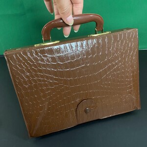 May include: A brown briefcase with a crocodile skin effect. It features a brown handle and gold-coloured metal clasps. The front flap is secured with a small, round clasp. The briefcase is rectangular.