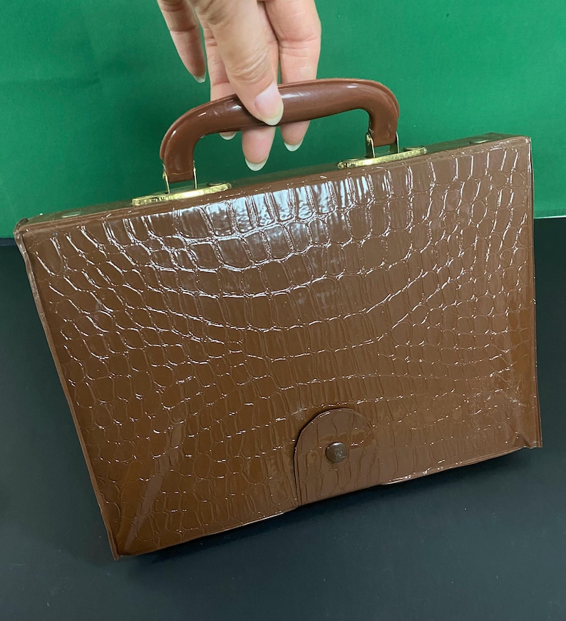 May include: A brown briefcase with a crocodile skin effect. It features a brown handle and gold-coloured metal clasps. The front flap is secured with a small, round clasp. The briefcase is rectangular.