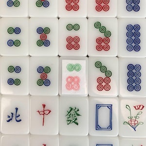 May include: A close-up shot of white Mahjong tiles with various symbols and characters in red, green, and blue. The tiles feature circles and Chinese characters. The tiles are arranged in a grid pattern.