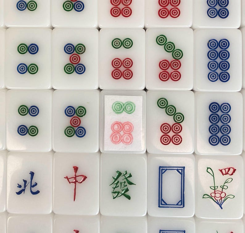 May include: A close-up shot of white Mahjong tiles with various symbols and characters in red, green, and blue. The tiles feature circles and Chinese characters. The tiles are arranged in a grid pattern.
