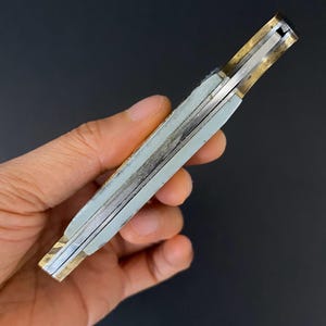 May include: A closed pocket knife with a light gray handle and brass-colored accents. The knife's blade is silver, and the handle appears to be made of a composite material. The knife is held in a hand against a dark background.