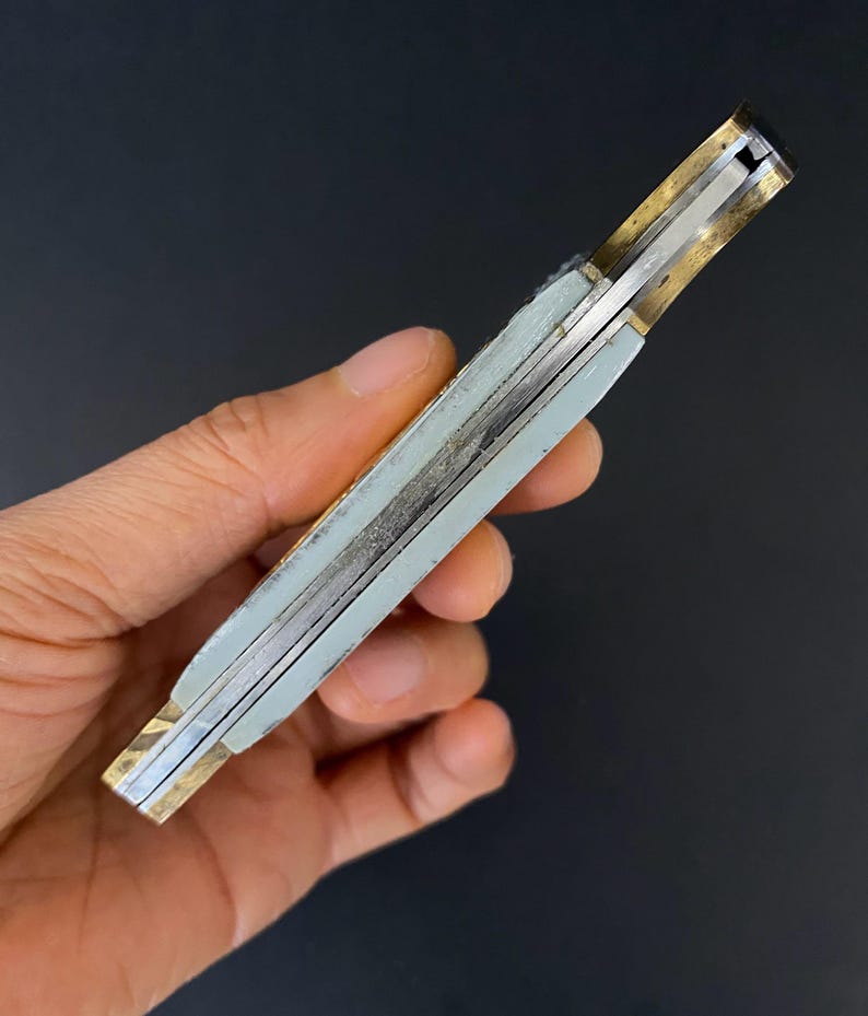 May include: A closed pocket knife with a light gray handle and brass-colored accents. The knife's blade is silver, and the handle appears to be made of a composite material. The knife is held in a hand against a dark background.