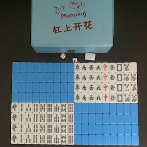 May include: A Mahjong set featuring blue tiles and a light blue box. The box is printed with the words "I Mahjong" and Chinese characters. The set includes dice and white tiles with Chinese characters and symbols, perfect for the traditional game.