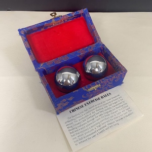 May include: Two shiny, chrome-like Chinese exercise balls rest inside a blue box with a red velvet interior. The box has a gold floral pattern and a small metal clasp. A white paper with text about the balls is also visible.