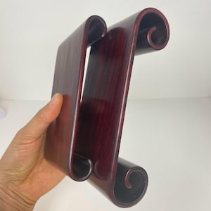 May include: A dark burgundy wooden display stand with a unique scroll design. The stand has a rectangular base and two curved, scroll-shaped supports. The wood grain is visible, and the finish is glossy.