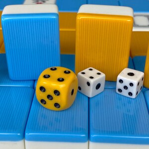 Puede incluir: Primer plano de fichas y dados de Mahjong. Las fichas son azules y amarillas con tapas blancas. Un dado amarillo con puntos negros se encuentra junto a dos dados blancos con puntos negros. Las piezas del juego est&aacute;n sobre una superficie azul y blanca.