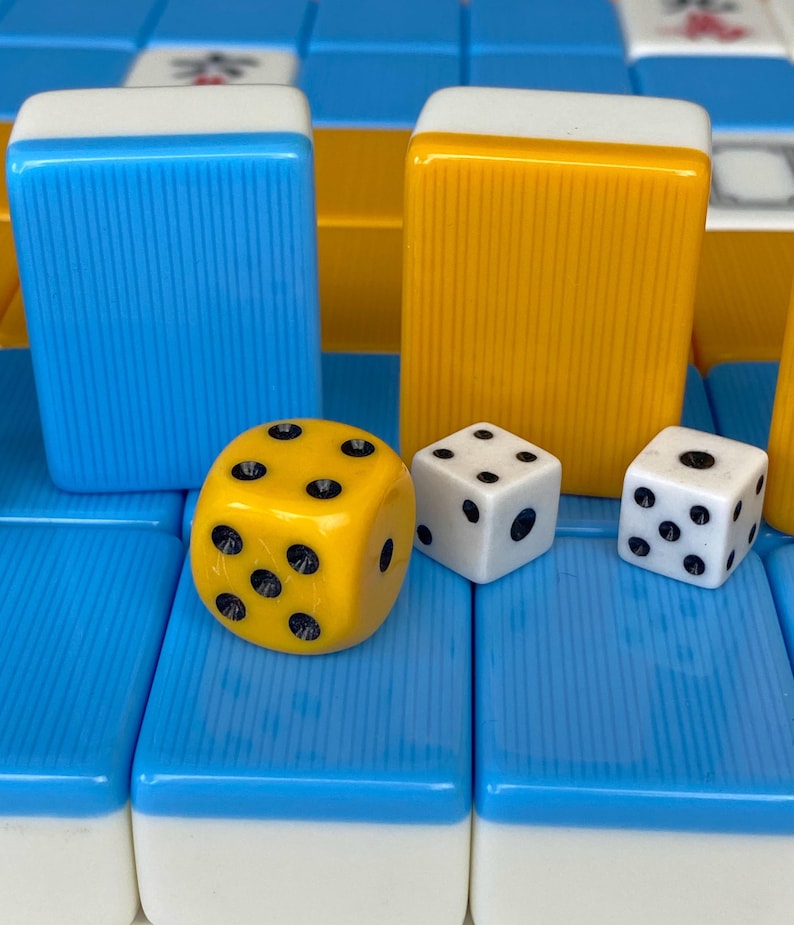 Puede incluir: Primer plano de fichas y dados de Mahjong. Las fichas son azules y amarillas con tapas blancas. Un dado amarillo con puntos negros se encuentra junto a dos dados blancos con puntos negros. Las piezas del juego est&aacute;n sobre una superficie azul y blanca.