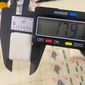 May include: A digital caliper measuring a white, rectangular Mahjong tile. The tile measures 1.79 cm in width. The caliper has an LCD screen displaying the measurement in mm and inches. Other Mahjong tiles are visible in the background.
