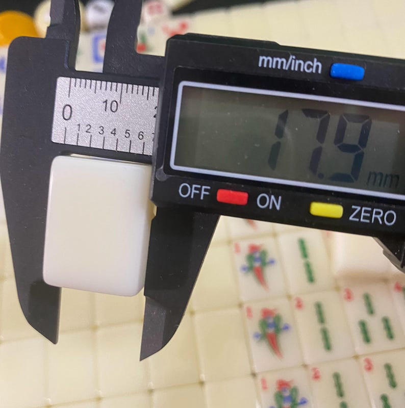 May include: A digital caliper measuring a white, rectangular Mahjong tile. The tile measures 1.79 cm in width. The caliper has an LCD screen displaying the measurement in mm and inches. Other Mahjong tiles are visible in the background.