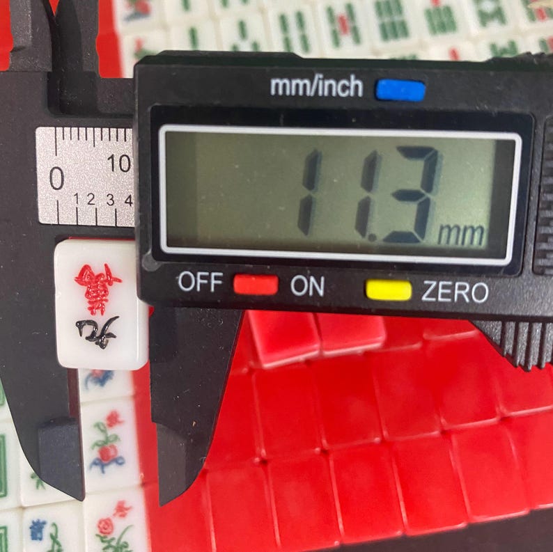 Puede incluir: Una ficha de Mahjong blanca con s&iacute;mbolos rojos y negros se mide con un calibrador digital. El calibrador muestra 11,3 mm. Hay fichas rojas visibles en el fondo.