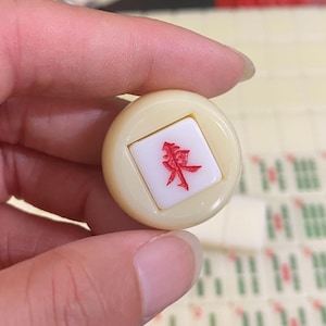 May include: A cream-coloured Mahjong tile with a red Chinese character is held between fingers. Other tiles are visible in the background. The character is in a square inset within the tile.