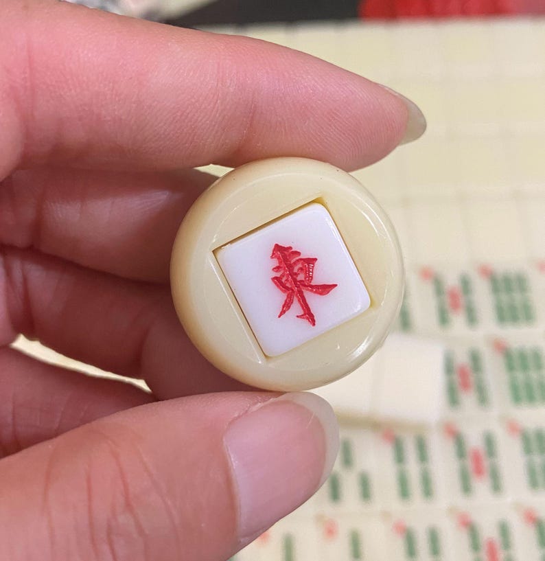 May include: A cream-coloured Mahjong tile with a red Chinese character is held between fingers. Other tiles are visible in the background. The character is in a square inset within the tile.