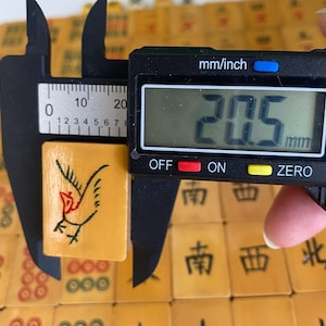 May include: A close-up of a vintage mahjong tile, measuring 20.5 mm. The tile is a light tan color with a black and red bird illustration. The tile is being measured with a digital caliper. Other mahjong tiles are in the background.