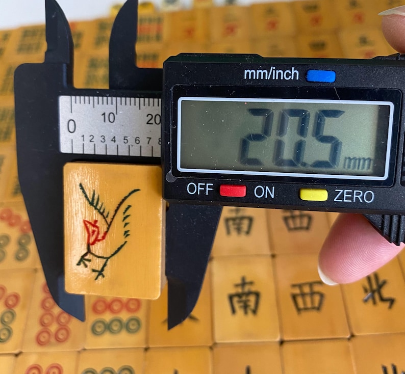 May include: A close-up of a vintage mahjong tile, measuring 20.5 mm. The tile is a light tan color with a black and red bird illustration. The tile is being measured with a digital caliper. Other mahjong tiles are in the background.