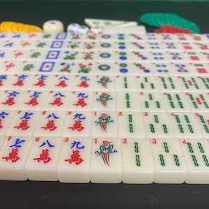May include: A full Mahjong set with cream-coloured tiles, displaying red, green, and blue symbols and numbers. Includes dice, counters, and racks. The tiles are arranged on a green surface, ready for play.