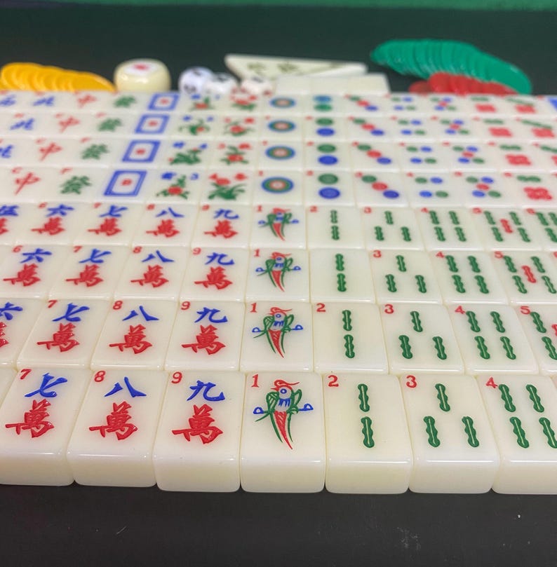 May include: A full Mahjong set with cream-coloured tiles, displaying red, green, and blue symbols and numbers. Includes dice, counters, and racks. The tiles are arranged on a green surface, ready for play.