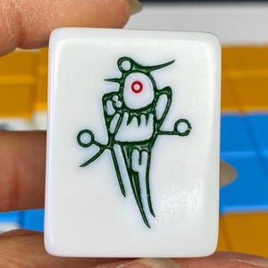 Puede incluir: Una ficha de Mahjong blanca con un dise&ntilde;o gr&aacute;fico verde. El dise&ntilde;o incluye una forma central en forma de ojo con un centro rojo, rodeada de intrincadas l&iacute;neas y formas verdes. La ficha es sostenida por una mano.