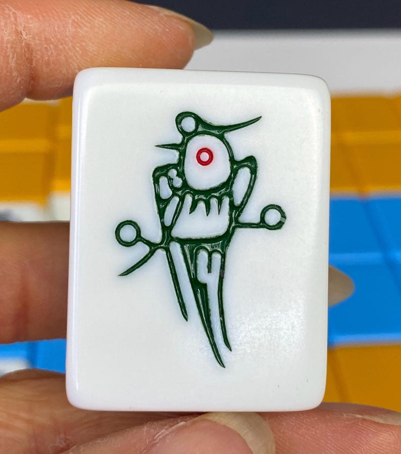 Puede incluir: Una ficha de Mahjong blanca con un dise&ntilde;o gr&aacute;fico verde. El dise&ntilde;o incluye una forma central en forma de ojo con un centro rojo, rodeada de intrincadas l&iacute;neas y formas verdes. La ficha es sostenida por una mano.