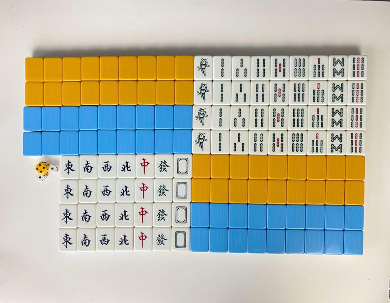 Puede incluir: Un juego de Mahjong completo con fichas dispuestas en filas. Las fichas son blancas con s&iacute;mbolos negros y rojos. El juego incluye fichas amarillas y azules, y un par de dados. El juego est&aacute; sobre una superficie blanca.