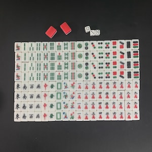 Puede incluir: Un juego completo de Mahjong con fichas blancas con caracteres y s&iacute;mbolos chinos, dados rojos y dos soportes rojos para fichas. Las fichas est&aacute;n dispuestas sobre una superficie negra.