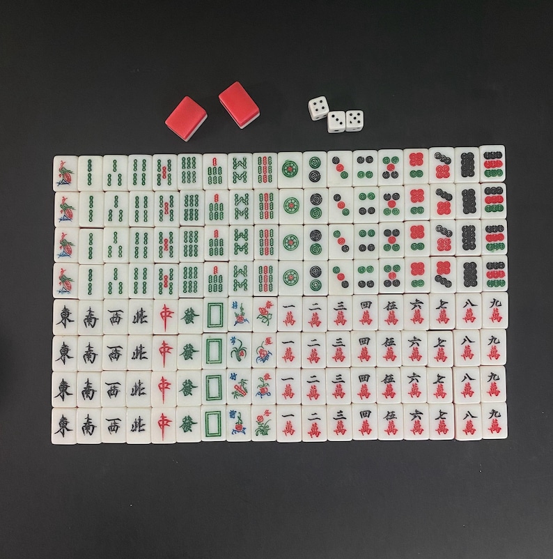 Puede incluir: Un juego completo de Mahjong con fichas blancas con caracteres y s&iacute;mbolos chinos, dados rojos y dos soportes rojos para fichas. Las fichas est&aacute;n dispuestas sobre una superficie negra.