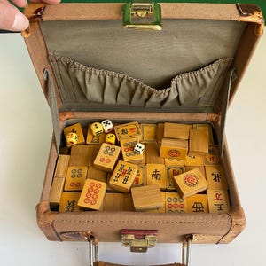 May include: Open vintage brown suitcase filled with wooden Mahjong tiles and dice. The tiles have red and black markings. The suitcase has a green clasp and a brown handle.
