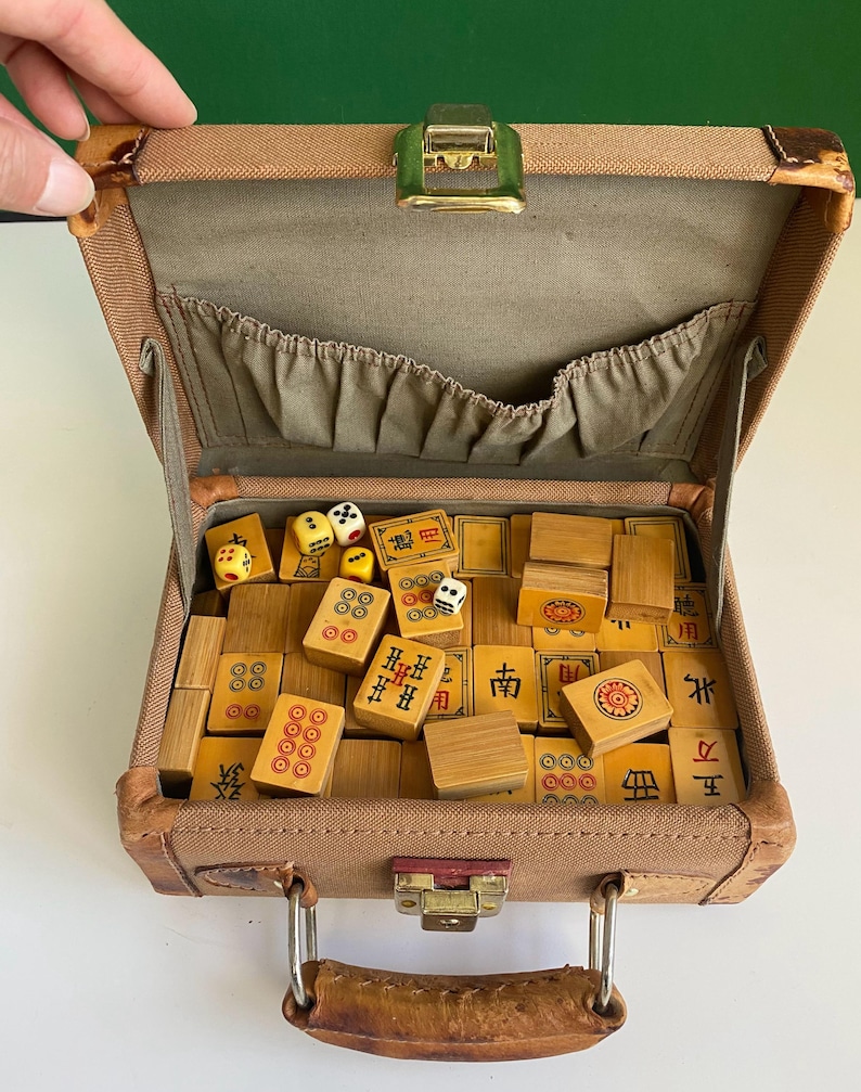 May include: Open vintage brown suitcase filled with wooden Mahjong tiles and dice. The tiles have red and black markings. The suitcase has a green clasp and a brown handle.