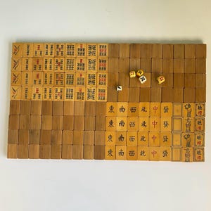 May include: A vintage Mahjong set with wooden tiles and dice. The tiles feature Chinese characters and symbols in red and black ink. The set is arranged on a white surface, showcasing the game's components.
