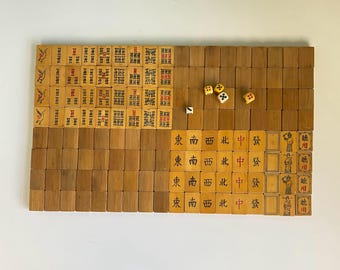May include: A vintage Mahjong set with wooden tiles and dice. The tiles feature Chinese characters and symbols in red and black ink. The set is arranged on a white surface, showcasing the game's components.