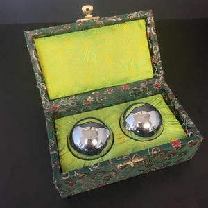 Può includere: Un set di due sfere metalliche lucide, cromate, all'interno di una scatola foderata in raso verde. La scatola è verde scuro con un motivo floreale e una chiusura dorata. L'interno è verde brillante.